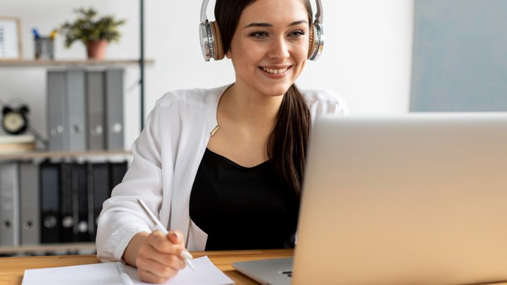 A person wearing headphones following a tutorial on a laptop and writing down the steps